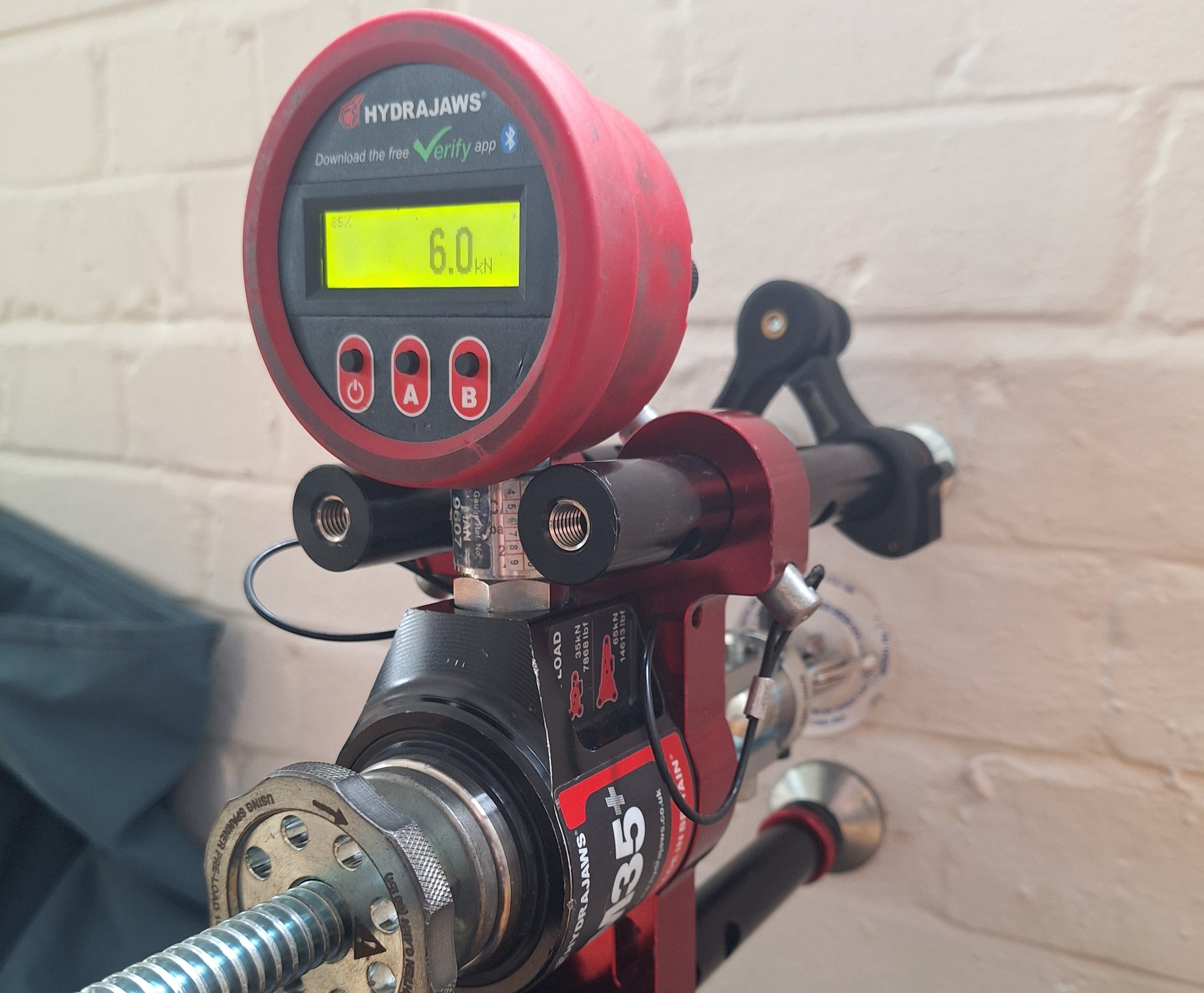 Hydrajaws M35+ anchor load testing rig reading 6.0 kN mounted against a white brick wall